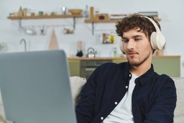 Şık kıvırcık saçlı bir adam evde rahat rahat otururken kulaklıkla müzik dinler..
