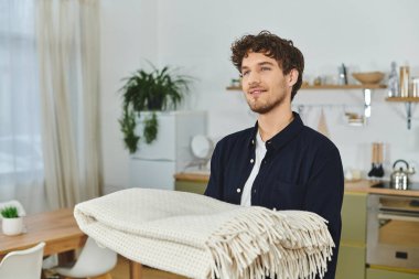 Tişörtlü kıvırcık saçlı adam elinde bir battaniye, canlı yaşam alanında gülümsüyor..