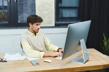 Kıvırcık saçlı süveter giyen adam çağdaş ofis ortamında bilgisayar görevlerine odaklanıyor.