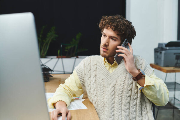 Curly-haired young man in a sweater vest communicates on the phone while working at his desk.