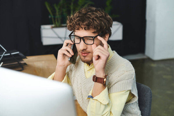 Young man in a sweater vest communicates on a phone while working in a modern office setting.