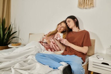 İki genç insan, rahat bir yatakta oturmuş sıcak içeceklerin tadını çıkarırken huzurlu bir anı paylaşıyor..