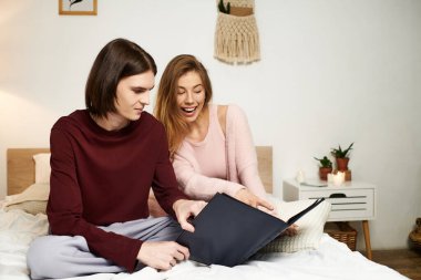 A stylish couple laughs together while looking through a scrapbook in their cozy bedroom. clipart