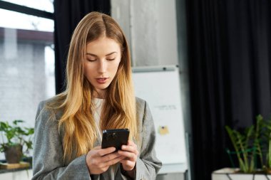 Şık bir genç kadın, çağdaş bir ofis ortamında dururken telefonuna odaklanır..