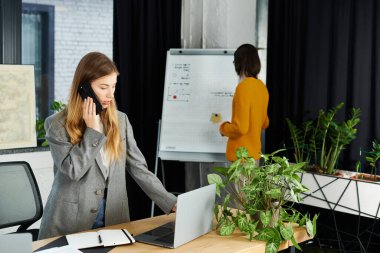 Şık giyinmiş genç bir kadın, meslektaşı sunuma hazırlanırken bir telefon görüşmesine odaklanıyor..