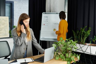 Young woman in chic attire focuses on a phone call as her colleague prepares for a presentation. clipart