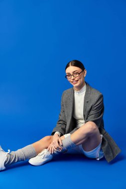 A beautiful young woman expresses emotion while seated stylishly on a bright blue background. clipart