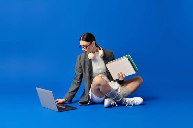 Young beautiful woman with headphones focuses on her laptop, surrounded by colorful notebooks. clipart