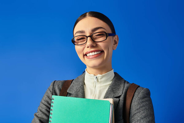 Радостная девушка в очках обнимает свой блокнот в уютной обстановке.