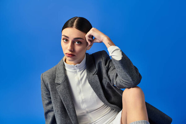 Молодая женщина выражает глубокие эмоции, сидя изящно, в окружении смелого синего цвета.