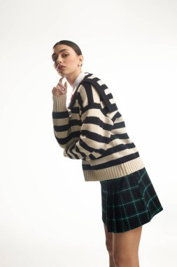 Emotional young woman with a playful expression wearing a striped sweater and tartan skirt, indoors. clipart