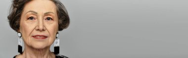 60 'lı yaşlarda güzel bir kadın zarif bir şekilde poz verir. Tarafsız bir arka plan, afiş karşısında.