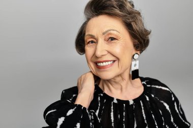 60 'lı yaşlarda şık bir kadın, gri bir arka plana karşı güvenle parlar..