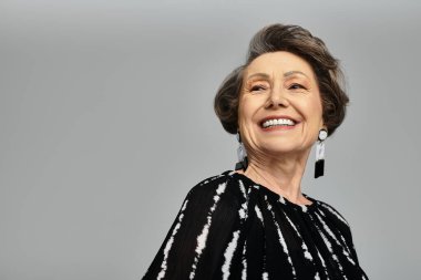 Olgun bir kadın şık giyinerek parıldar. Gri bir arka plana karşı özgüven ve zarafet gösterir..
