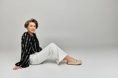 60 'lı yaşlarda güzel bir kadın, parlak gri bir arka plana karşı şık giyiniyor..