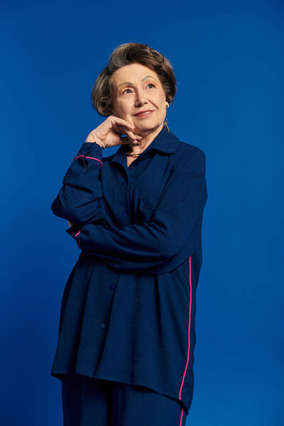 A stylish woman in her 60s poses confidently in fashionable clothing with a blue backdrop.