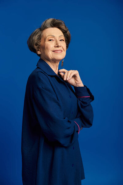 A confident woman in her 60s elegantly poses in fashionable attire, radiating beauty.