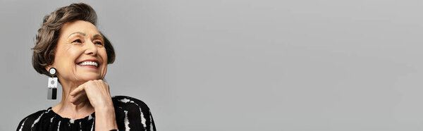 A beautiful woman in her 60s confidently poses in stylish attire against a grey backdrop, banner