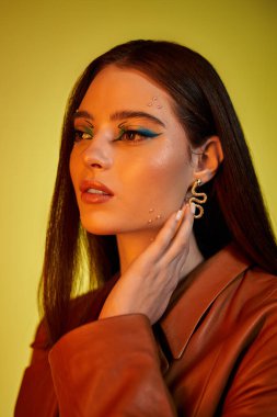 Genç bir kadın zarif bir pozla göz kamaştırıcı bir makyaj ve kıyafetle canlı bir arkaplana karşı duruyor..