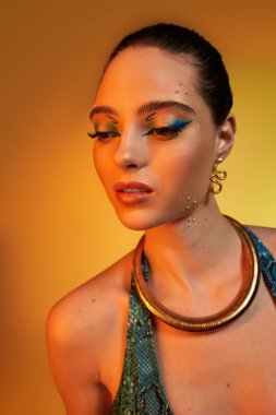 Genç bir kadın, sıcak bir arkaplana karşı şık yılan desenli kıyafetler ve kalın bir makyaj sergiliyor.
