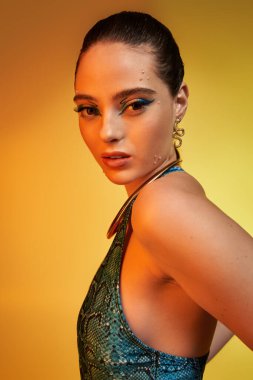 Yılan desenli elbiseli ve çarpıcı makyajlı genç bir kadın parlak bir arkaplanda poz veriyor.