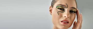 Beautiful young woman poses elegantly in a studio, displaying intricate snake makeup art, banner clipart