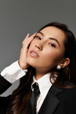 Genç bir kadın, gri arka planda uzun kahverengi saçlı, şık bir elbiseyle rahatça poz veriyor..