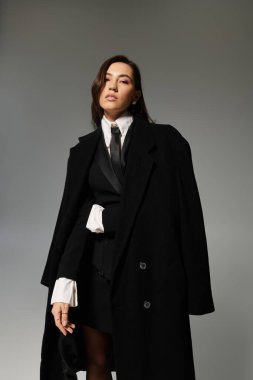 Uzun kahverengi saçlı, kendine güvenen genç bir kadın gri bir arkaplanda şık kıyafetler içinde poz veriyor..