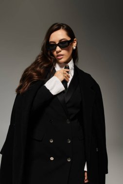 Uzun kahverengi saçlı güzel bir kadın gri bir arkaplana karşı şık bir kıyafet sergiliyor..