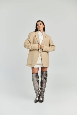 Genç bir kadın kendine güvenerek ayakta duruyor, şık, büyük bir spor ceket ve diz boyu metalik çizmeler modelliyor..