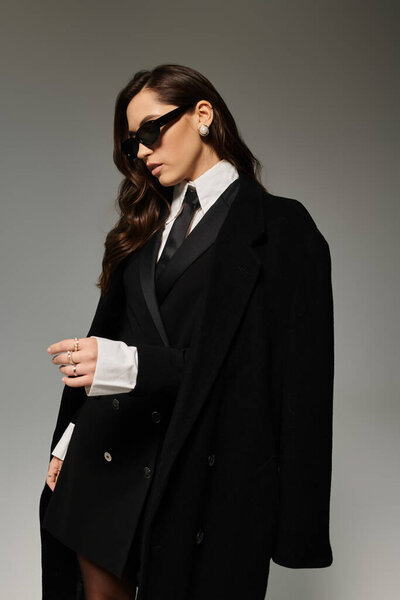 Красивая женщина с длинными каштановыми волосами стильно позирует в шикарном наряде на сером фоне.