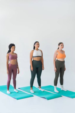 Üç güçlü kadın spor kıyafetleri içinde renkli minderlerin üzerinde dururken fitness yolculuklarını sergiliyorlar..