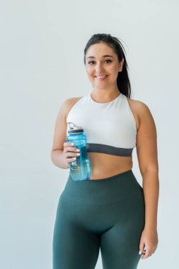 A fit woman smiles while holding a water bottle, ready for an energetic workout session. clipart