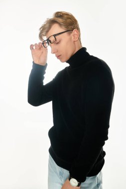 Siyah balıkçı yaka giyen genç bir adam gözlüğünü özgüvenli bir ifadeyle incelikle ayarlıyor..