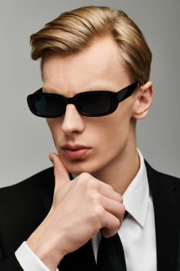 Güneş gözlüklü ve takım elbiseli genç adam tarafsız bir arkaplanda düşünceli bir duruş sergiliyor..