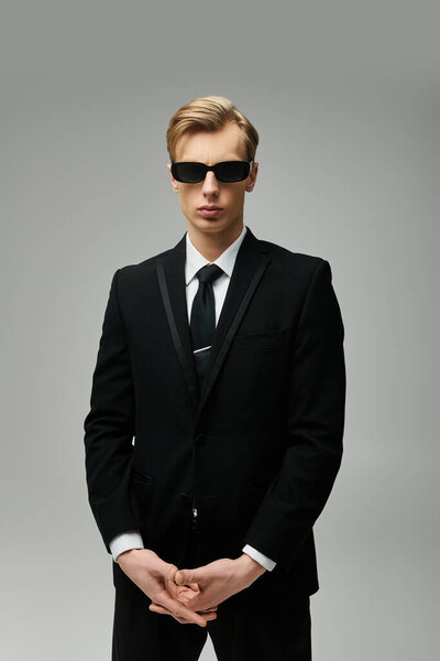 Stylish young man stands confidently with sunglasses and a sharp black suit, showcasing elegance.