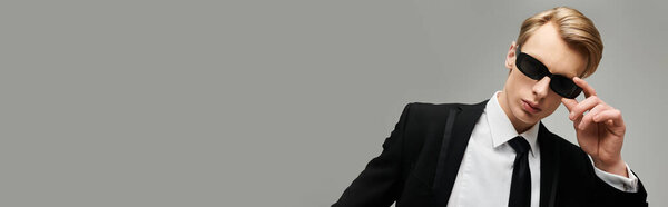 A young handsome man adjusts his sunglasses while wearing a classic black suit, showcasing elegance.