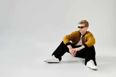 Güneş gözlüklü ve modaya uygun giysileri olan havalı bir genç adam minimalist bir arka planda oturuyor..