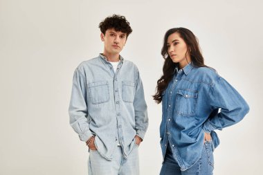 İki şık şahıs, temiz bir arkaplana karşı, modaya uygun kot kıyafetlerle rahatça poz veriyor..