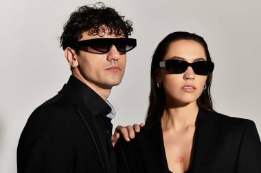 Genç bir çift, minimalist bir arkaplana karşı cesur güneş gözlüğü ve şık kıyafetler takarak ışıldıyor..