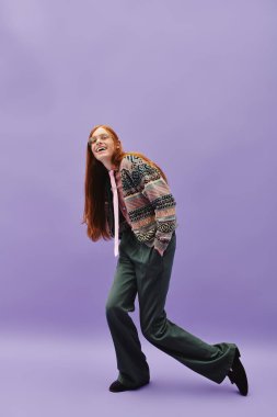 A young queer man with long red hair laughs while posing stylishly against a purple backdrop. clipart