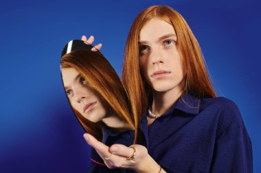 Young man with long red hair engages with a mirror against a striking blue background. clipart