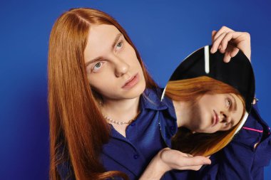 Genç eşcinsel bir adam mavi bir arkaplan önünde poz veriyor, elinde tarzını yansıtan bir ayna tutuyor..