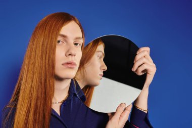 A stylish queer young man with red hair gazes thoughtfully into a mirror. clipart