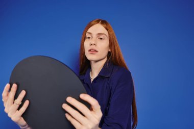 A stylish young queer man with long red hair thoughtfully gazes into a round mirror. clipart