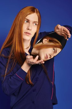 Modayı takip eden kızıl saçlı genç bir adam mavi bir arkaplan karşısında kendine has bir aynayla poz veriyor..