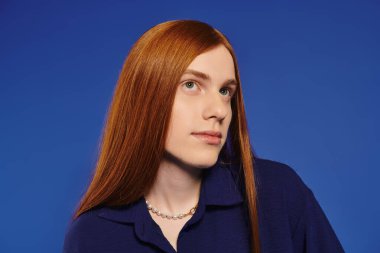 Şık, uzun kızıl saçlı genç bir adam parlak mavi bir arka plana karşı duruyor..