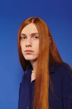 Uzun kızıl saçlı genç bir adam mavi arka planda kendine güvenen bir görünüm sergiliyor..