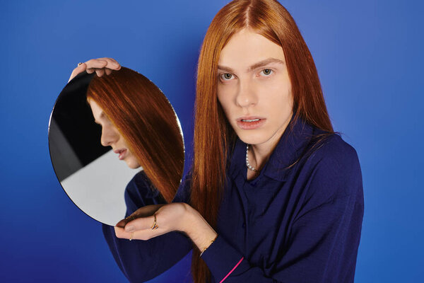 Handsome young man with long red hair holds a mirror, showcasing his bold, stylish essence.