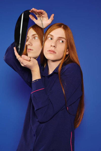 A young queer man with long red hair explores self expression, gazing into a mirror creatively held.